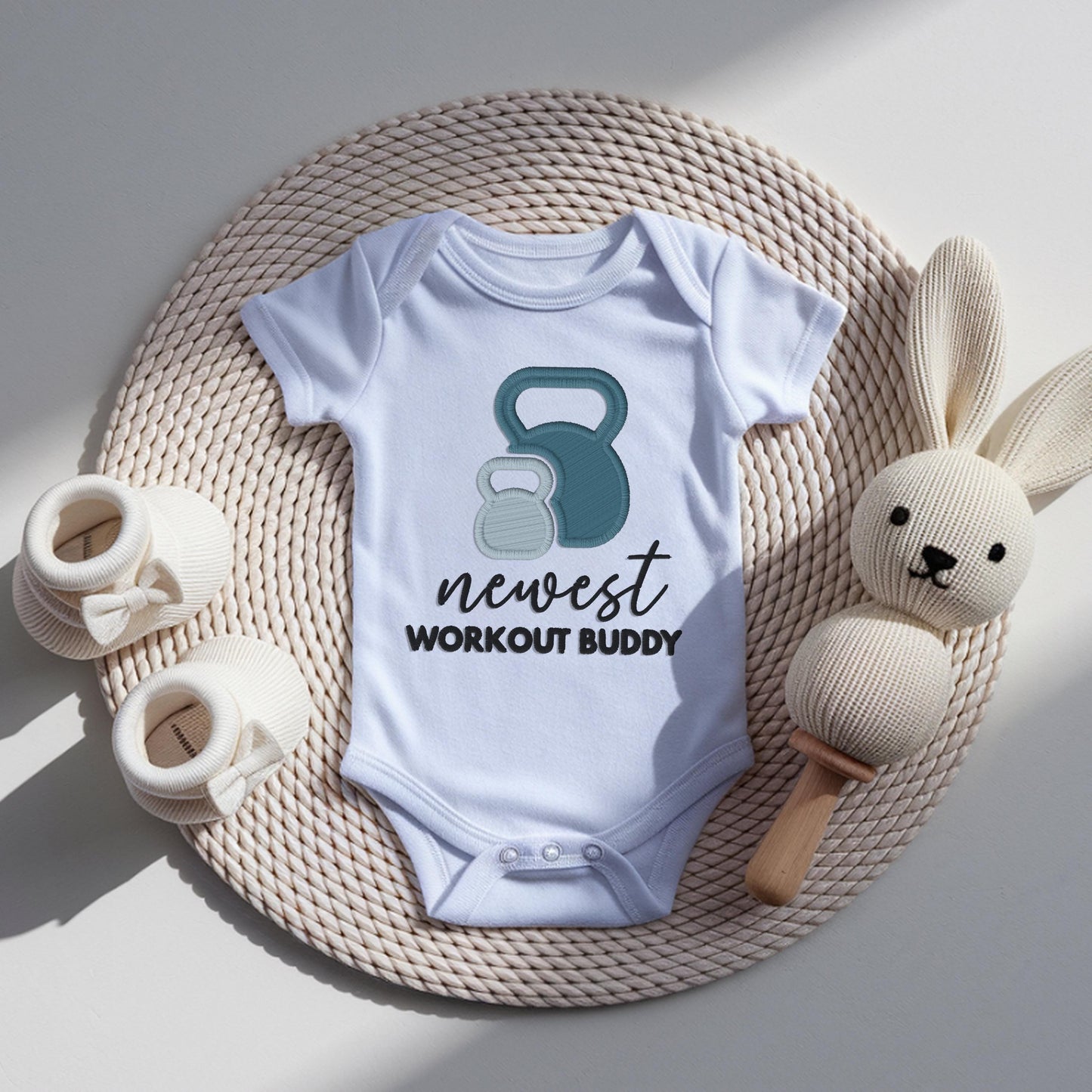 White baby bodysuit with embroidered kettlebell design and newest workout buddy text, fitness themed newborn outfit for pregnancy announcement.