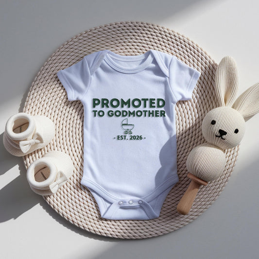 White baby bodysuit with raised embroidery reading “Promoted To Godmother” with small stroller icon and personalized EST year underneath in textured stitching.